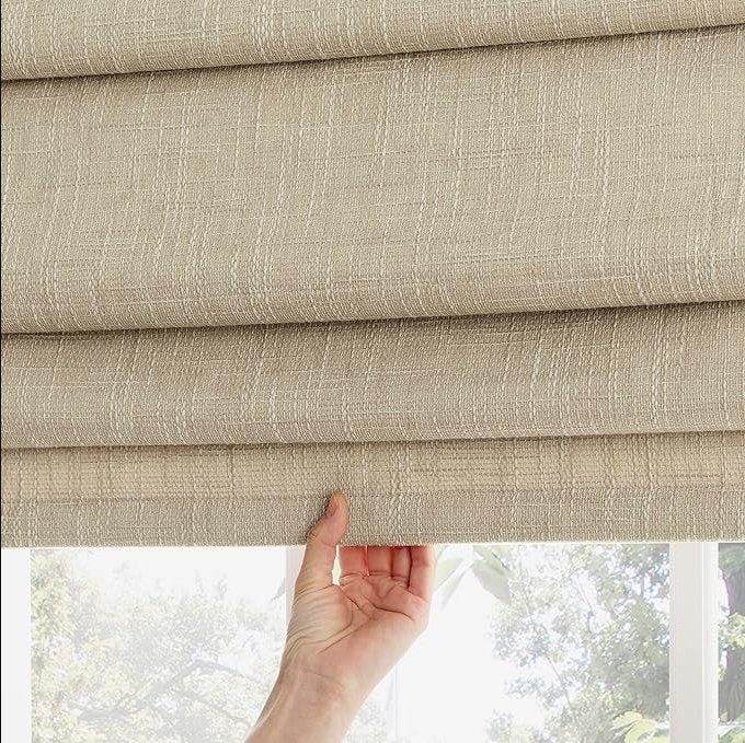 Hand adjusting textured beige blackout Roman shade with grid pattern, partially drawn to reveal outdoor greenery.