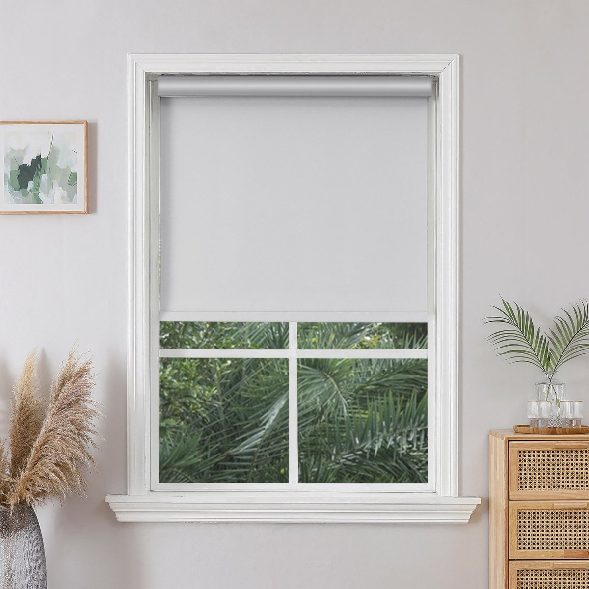 Bleach Joydeco light grey cordless blackout roller shade on a white trimmed window, showing lush green foliage outside.