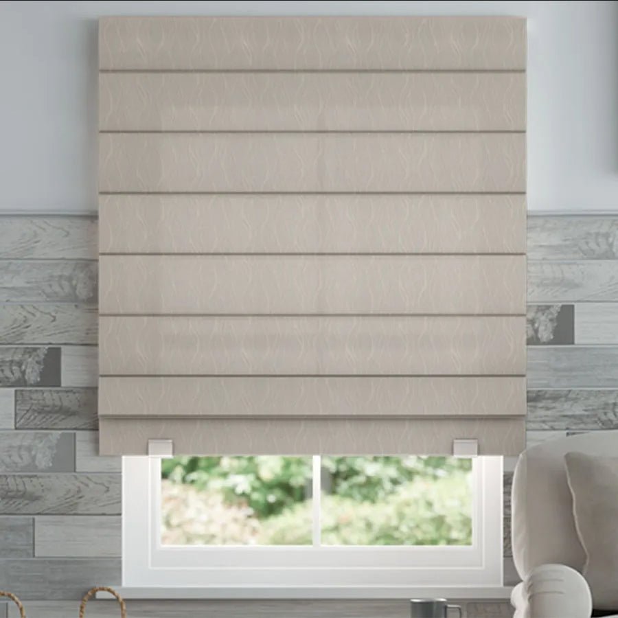 Joydeco beige blackout Roman shade with horizontal pleats, shown on a window with grey wood plank walls and greenery outside.
