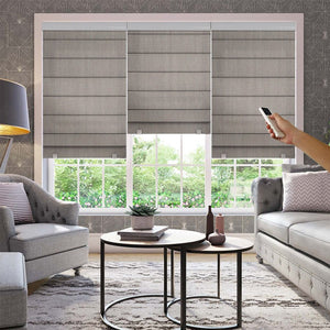 Hand holding remote to control three gray motorization blackout Roman shades on a window in a living room.