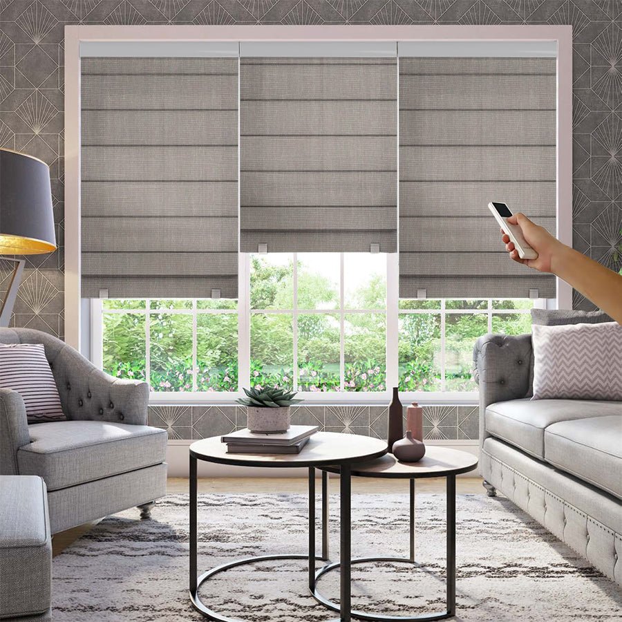 Hand holding remote to control three gray motorization blackout Roman shades on a window in a living room.