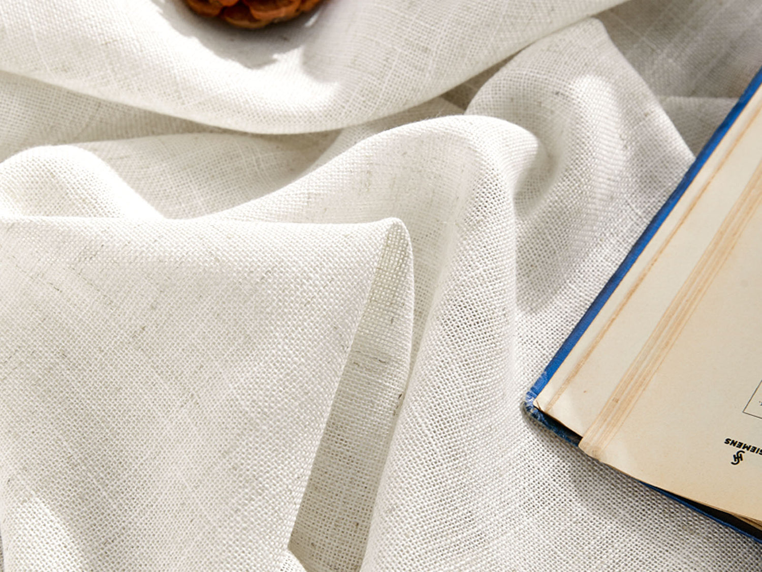 Close-up texture of wrinkled white linen fabric draped with an open book and a pine cone in soft natural light.