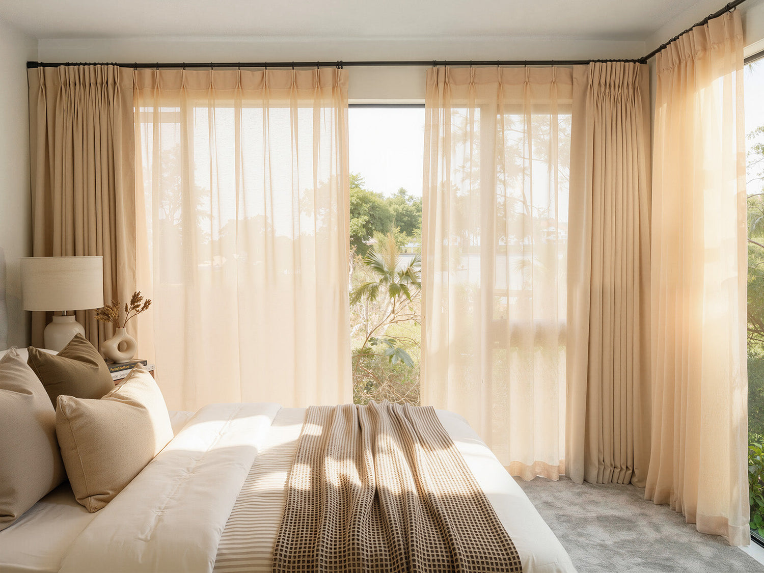 Sunlit bedroom with layered beige sheer and blackout curtains framing large corner windows