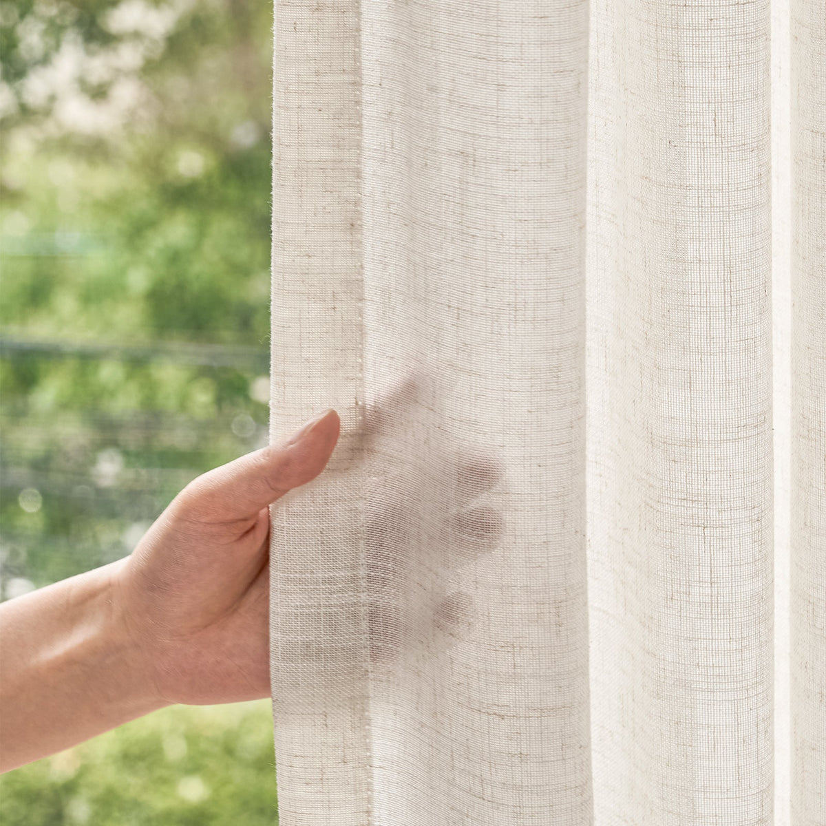 Close-up of hand touching off-white sheer curtain, showcasing fine woven texture and soft semi-sheer light diffusion with blurred green outdoor view.