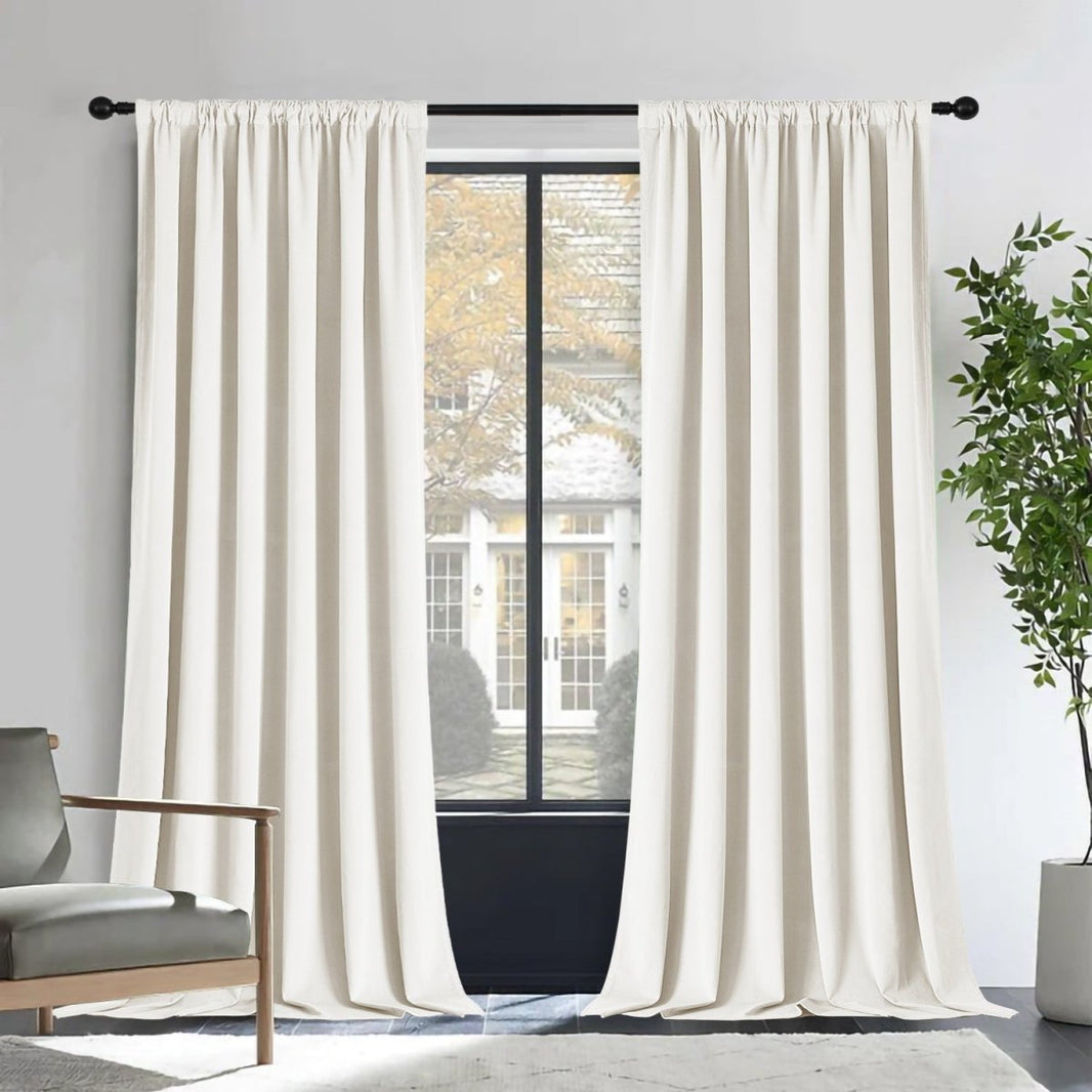 Two cream Joydeco velvet blackout curtains on black rod, room decor with armchair and plant.