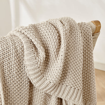 Close-up of beige 100% cotton chunky knit throw blanket showing intricate cable knit texture draped on wooden chair.