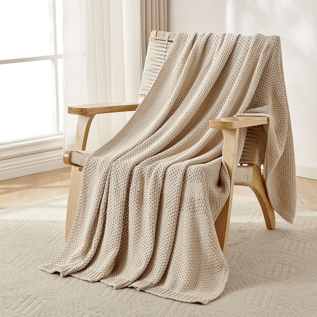 Beige chunky knit cotton throw blanket draped over a wooden armchair, showcasing farmhouse texture and coziness.