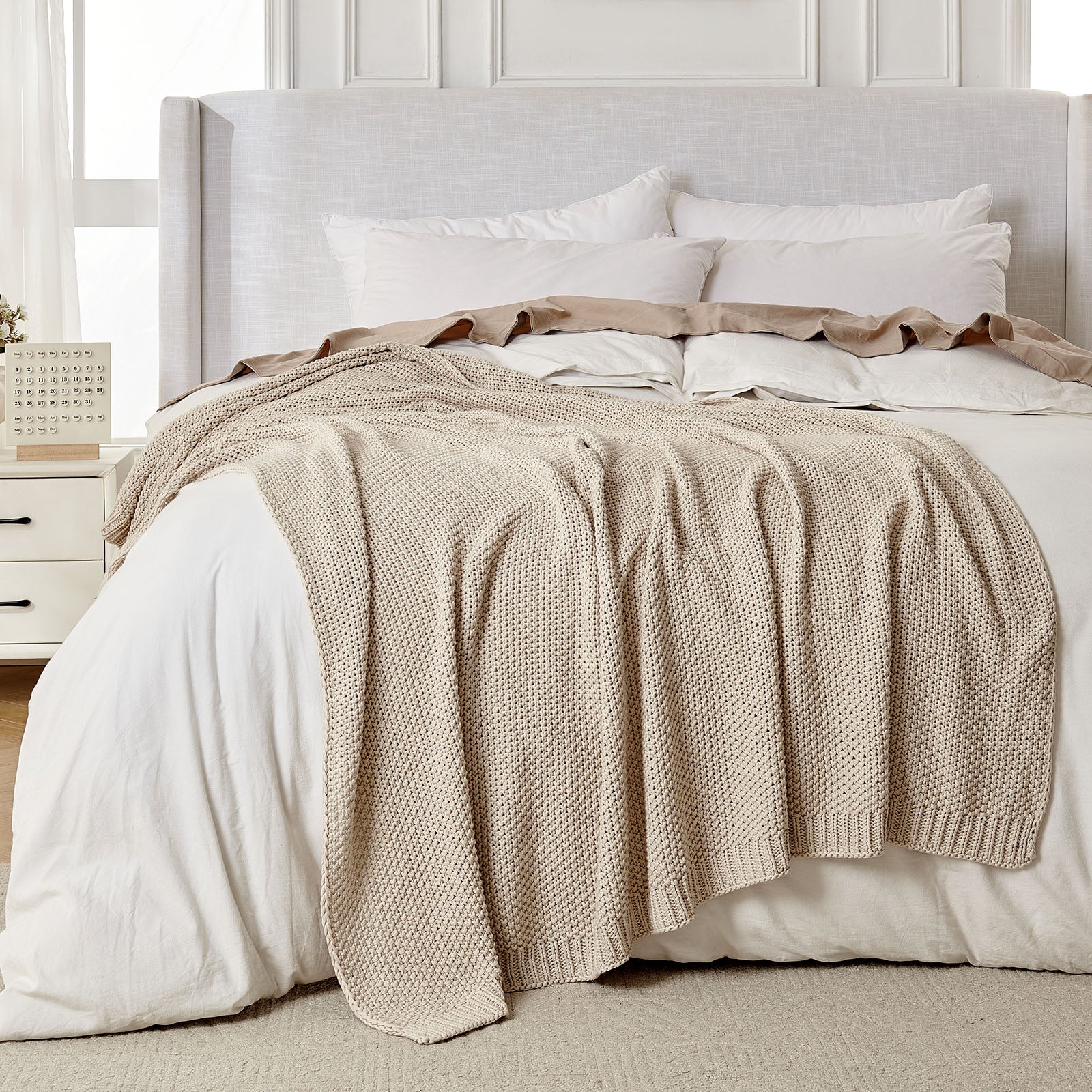 Light beige chunky knit 100% cotton throw blanket draped on a bed with white linens, showcasing texture and farmhouse style.