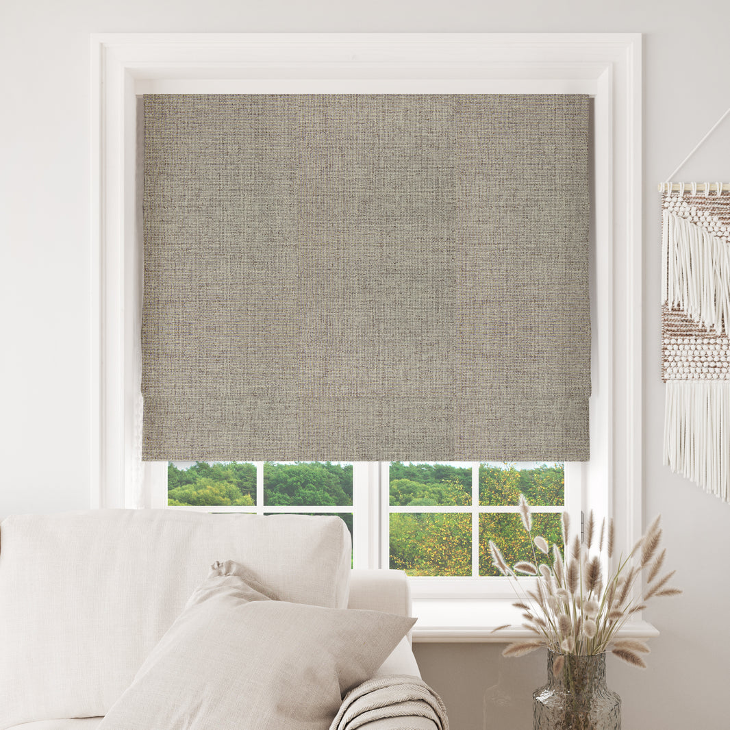 Joydeco greige blackout Roman shade with linen texture in white window, over sofa and greenery view.
