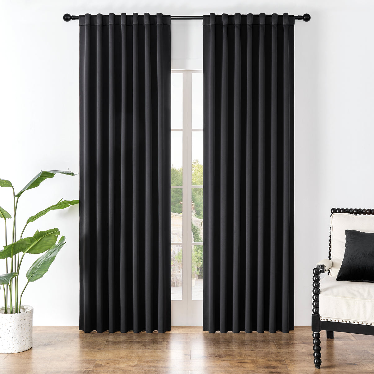 Joydeco black blackout curtains in a room setting with armchair and plant. Floor-length drapes with rod pocket and back tab.