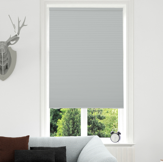 Light gray cordless blackout cellular shade, pleated honeycomb, on white frame, partially raised with trees outside.