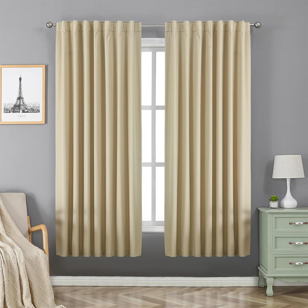Two beige Joydeco 100% blackout curtains with rod pocket and back tab hang over a window in a bedroom.