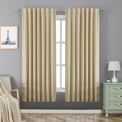 Two beige Joydeco 100% blackout curtains with rod pocket and back tab hang over a window in a bedroom.