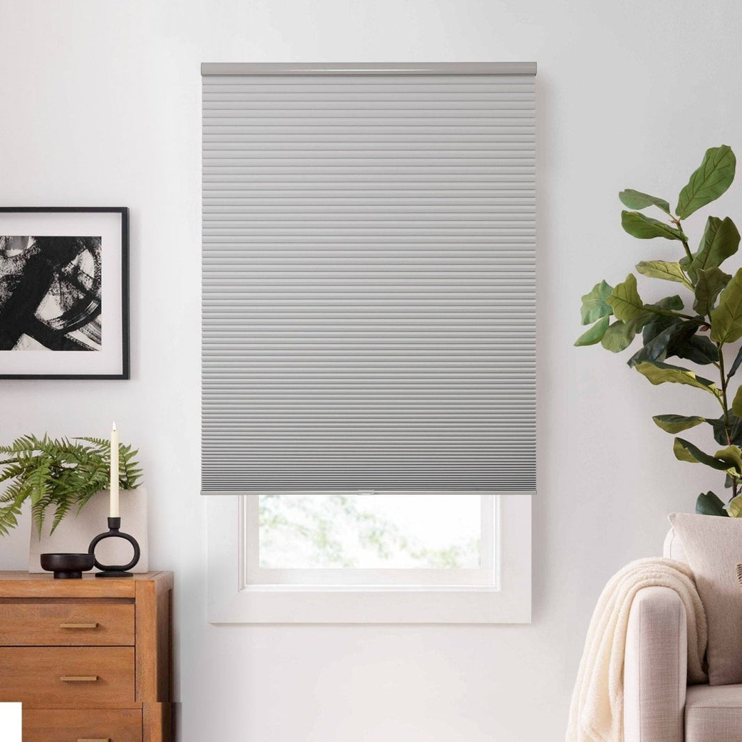 Light grey cordless blackout cellular shade on white window. Honeycomb texture visible. Home decor with dresser & plant.