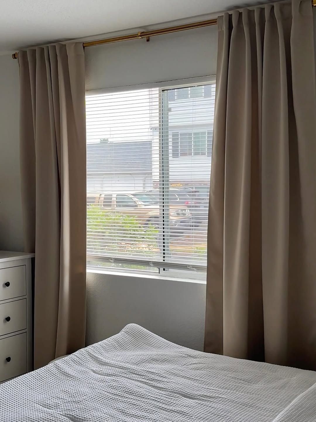 Joydeco beige blackout curtains on gold rod, blocking light from window with blinds, shown with a bed and duvet.