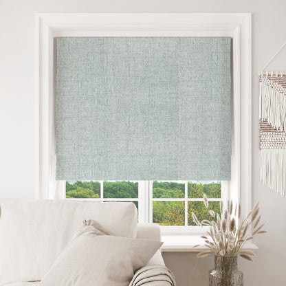 Celadon Joydeco blackout Roman shade, textured weave, installed on white window frame, view of trees.