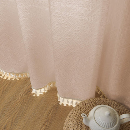 Close-up of light pink linen sheer curtain with geometric weave and cream tassel fringe detail, displayed on a woven pouf.