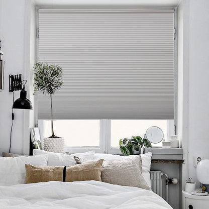 Light grey cordless blackout cellular shade on a window above a bed, showing pleated honeycomb design in a modern bedroom.