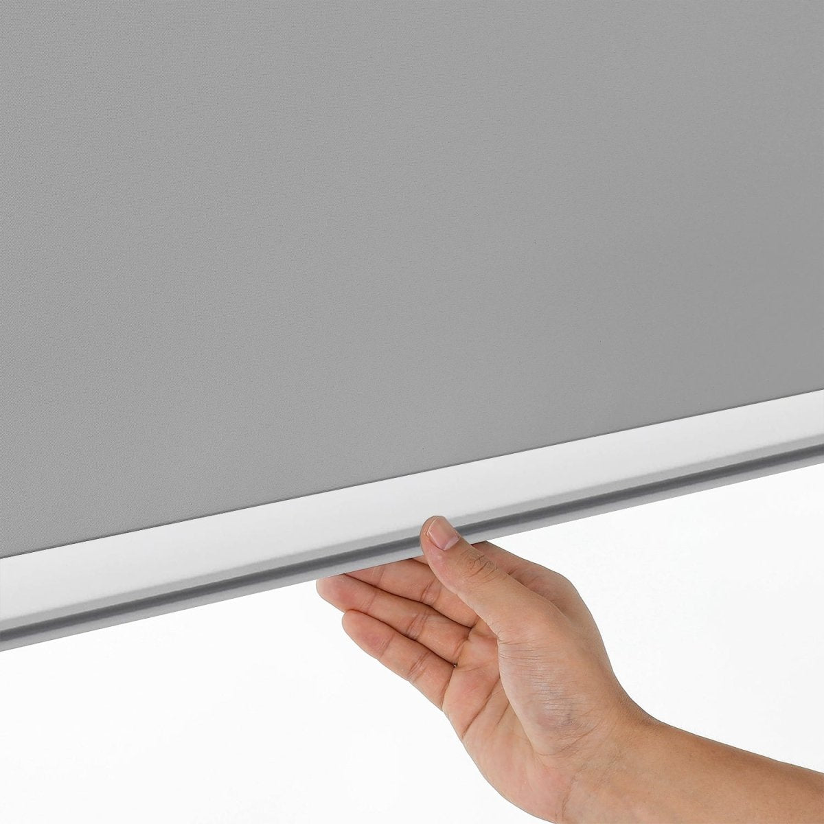 Close-up of a hand operating a light gray cordless blackout roller shade, showing the smooth fabric and aluminum bottom rod.