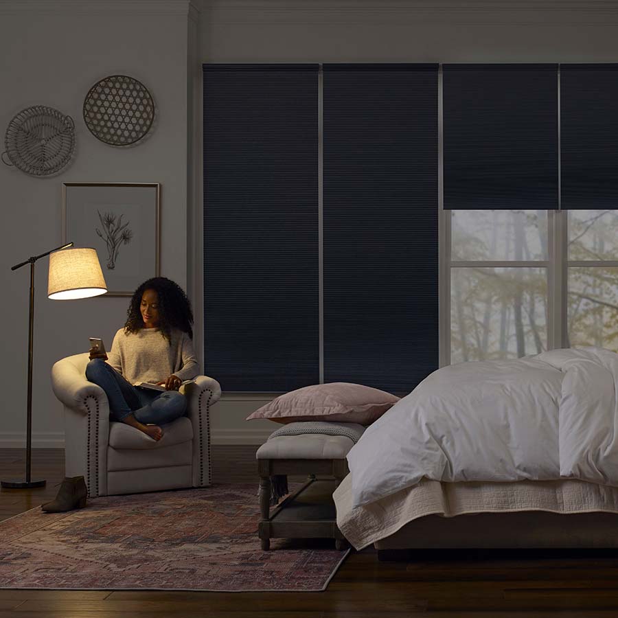 Joydeco blackout cellular shades in bedroom, woman relaxing by lamp, providing light control and privacy.